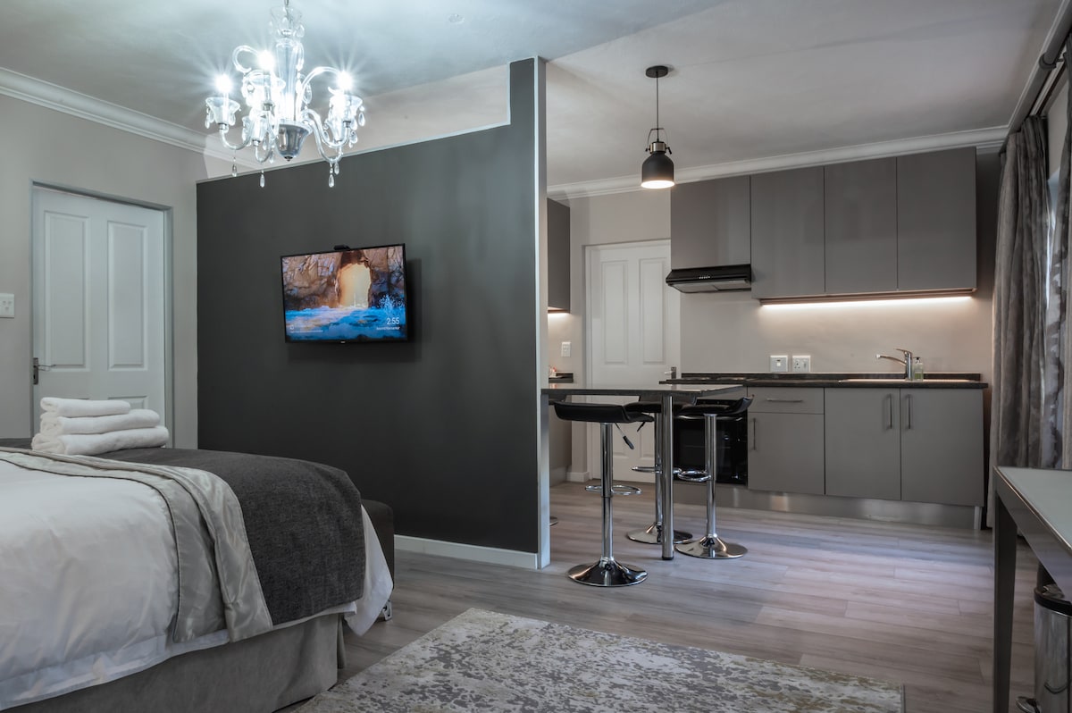 A well-appointed living space is illuminated by a decorative chandelier. A flat-screen TV is mounted on a grey wall, while a fully equipped kitchen features modern cabinetry. A dining area includes two sleek chairs and a round table, complemented by soft, neutral tones throughout the room.