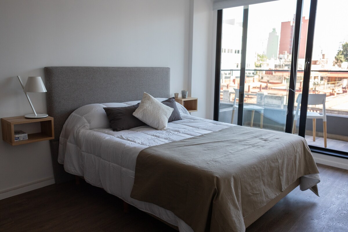 A modern bedroom features a spacious bed dressed in neutral tones with a decorative throw. Nightstands are positioned on either side, holding minimalistic lamps. Large glass doors flood the room with natural light, leading to a balcony with a view of the urban landscape.