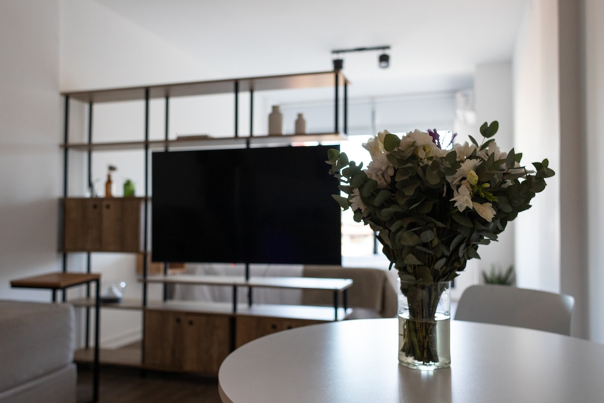 A modern living area features a sleek television positioned on a stylish shelving unit. A small table with a vase filled with greenery and delicate flowers is centered in the foreground, enhancing the contemporary design elements present in the space.