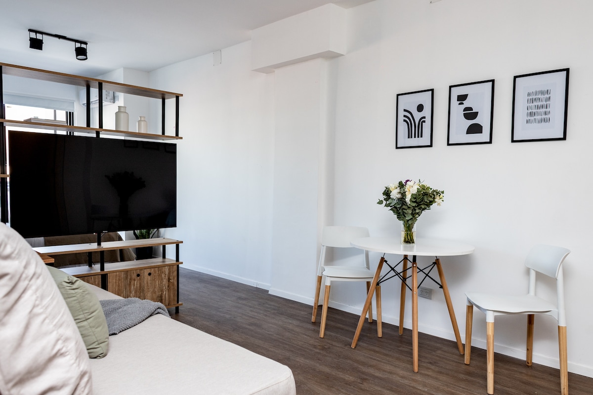 An open living space features a comfortable sofa and a round dining table surrounded by three chairs. A television is mounted on a wooden stand, while three framed artworks are displayed on the white wall, adding a touch of simplicity to the area.