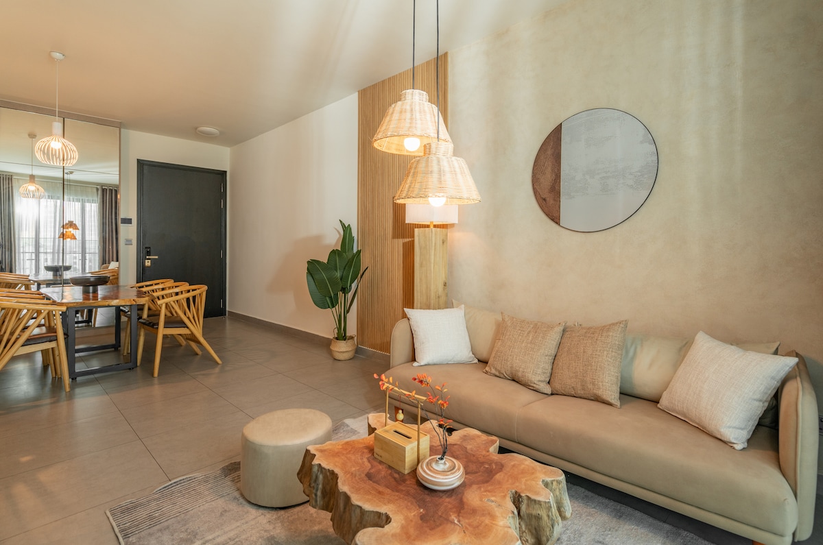A modern living area features a light-colored sofa accented by neutral pillows. A natural wood coffee table sits at the center, surrounded by a rug. Dual pendant lights hang above, illuminating the space. Nearby, a dining area with wooden chairs is visible.