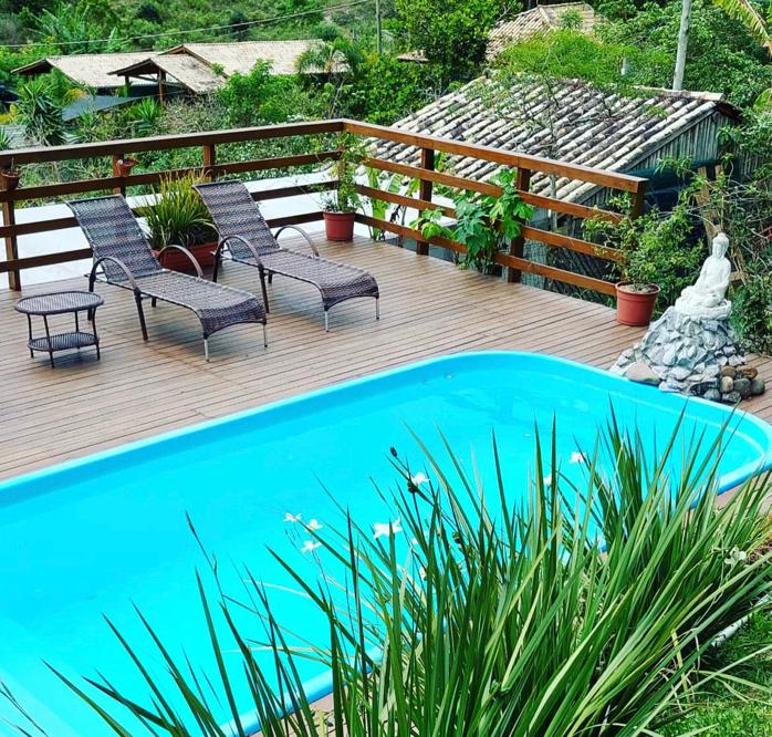 A serene outdoor area features a clear blue pool surrounded by wooden decking. Two lounge chairs are positioned thoughtfully beside the pool, and potted plants add green accents. A small sculpture of a seated figure is situated on a stone base, enhancing the tranquil environment.