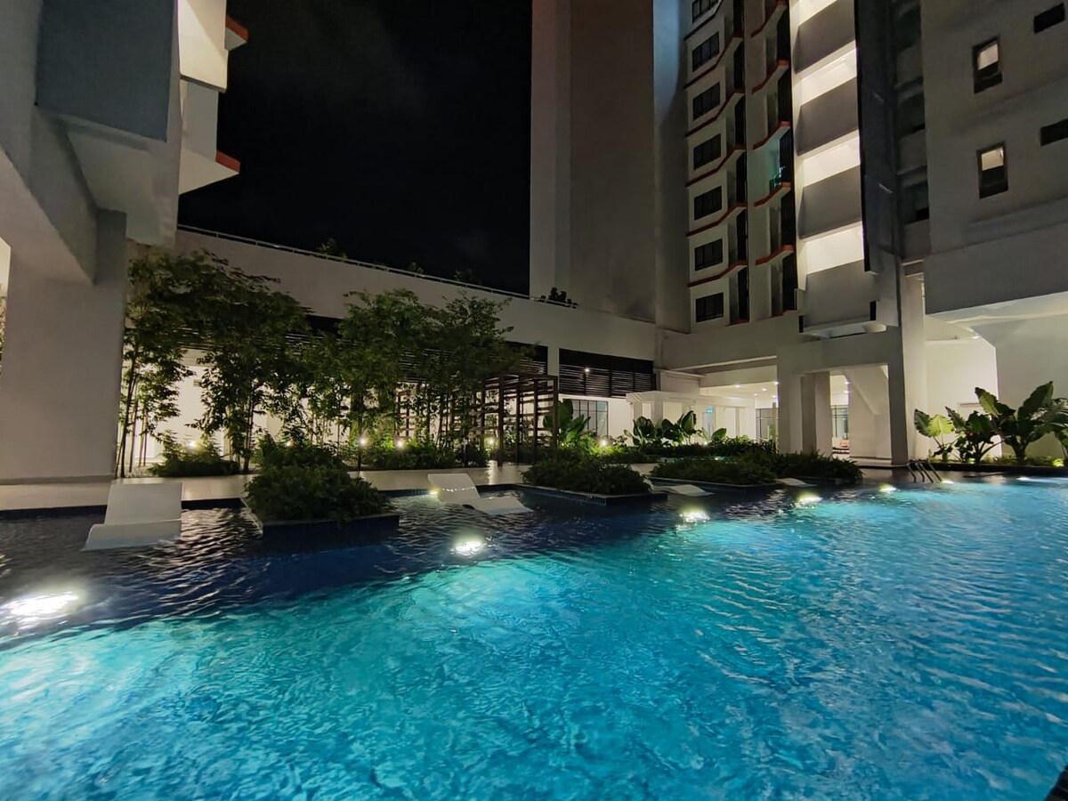 A serene swimming pool glimmers under the night sky, surrounded by lush greenery and softly illuminated landscaping. The water appears inviting and clear, with elegant lounge chairs positioned nearby, creating an ideal space for relaxation and comfort.