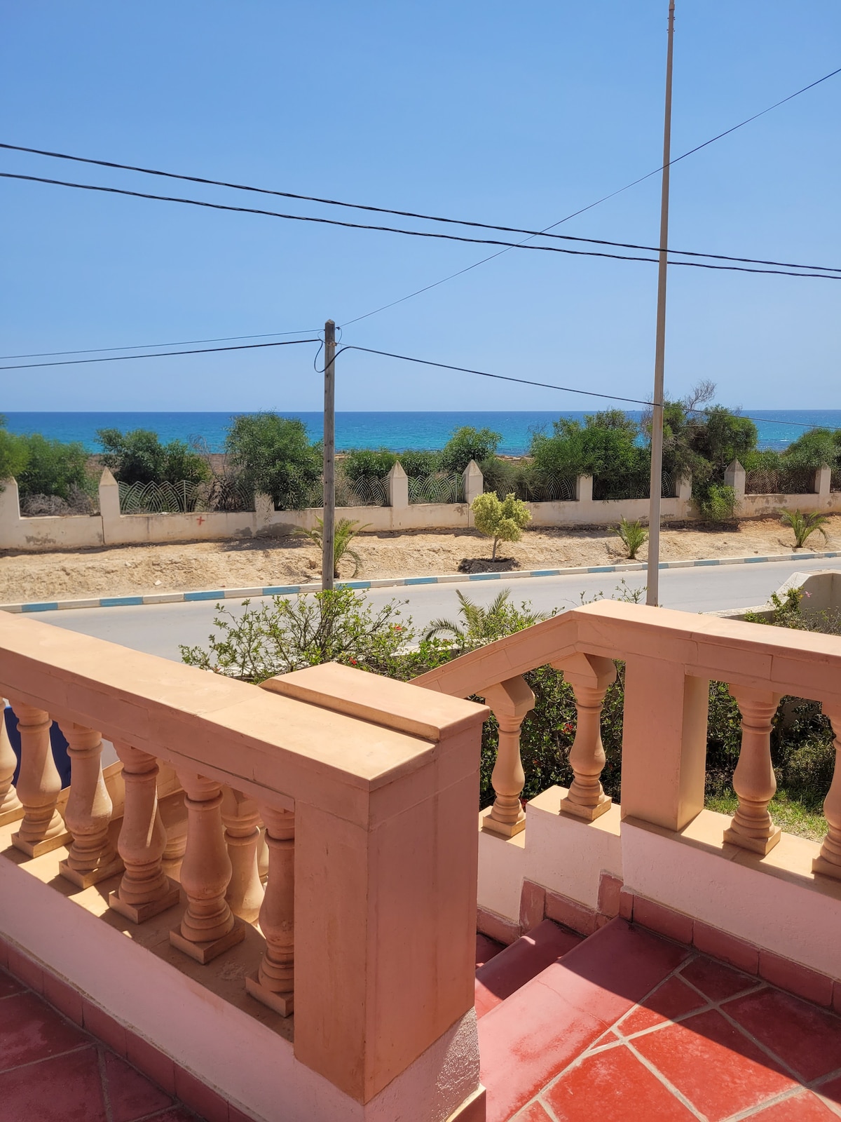 A view from the terrace showcases the serene sea, framed by lush greenery and the coastal road below. Clear blue skies complement the inviting atmosphere, while the balcony's decorative railing provides a charming focal point in the foreground.