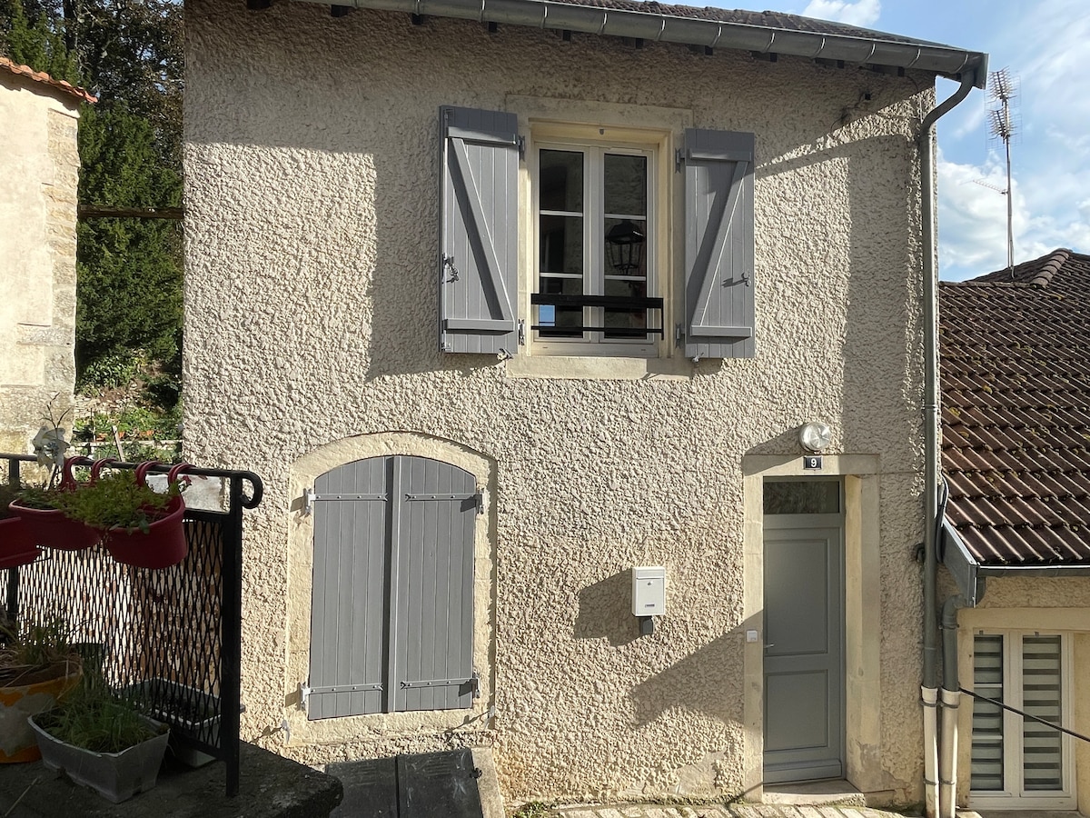 The exterior of the duplex apartment features a textured stone facade with gray shutters on the upper window. A wooden door is situated to the right, and a mailbox is mounted on the wall. Planter boxes with greenery can be seen on the left side.