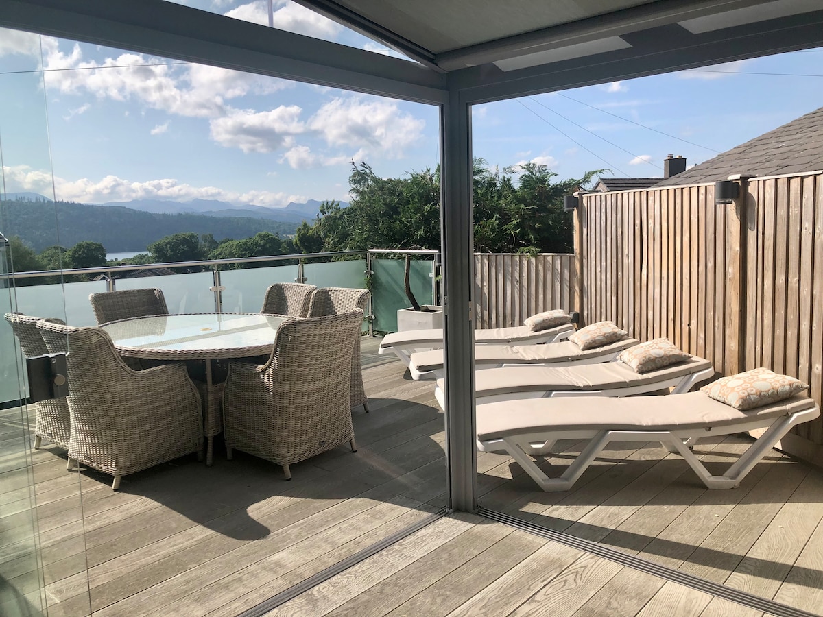 A spacious raised deck features a large dining table surrounded by six rattan chairs and several sun loungers. The area is enclosed by glass walls, offering uninterrupted views of the surrounding landscape and providing a sheltered environment for relaxation.