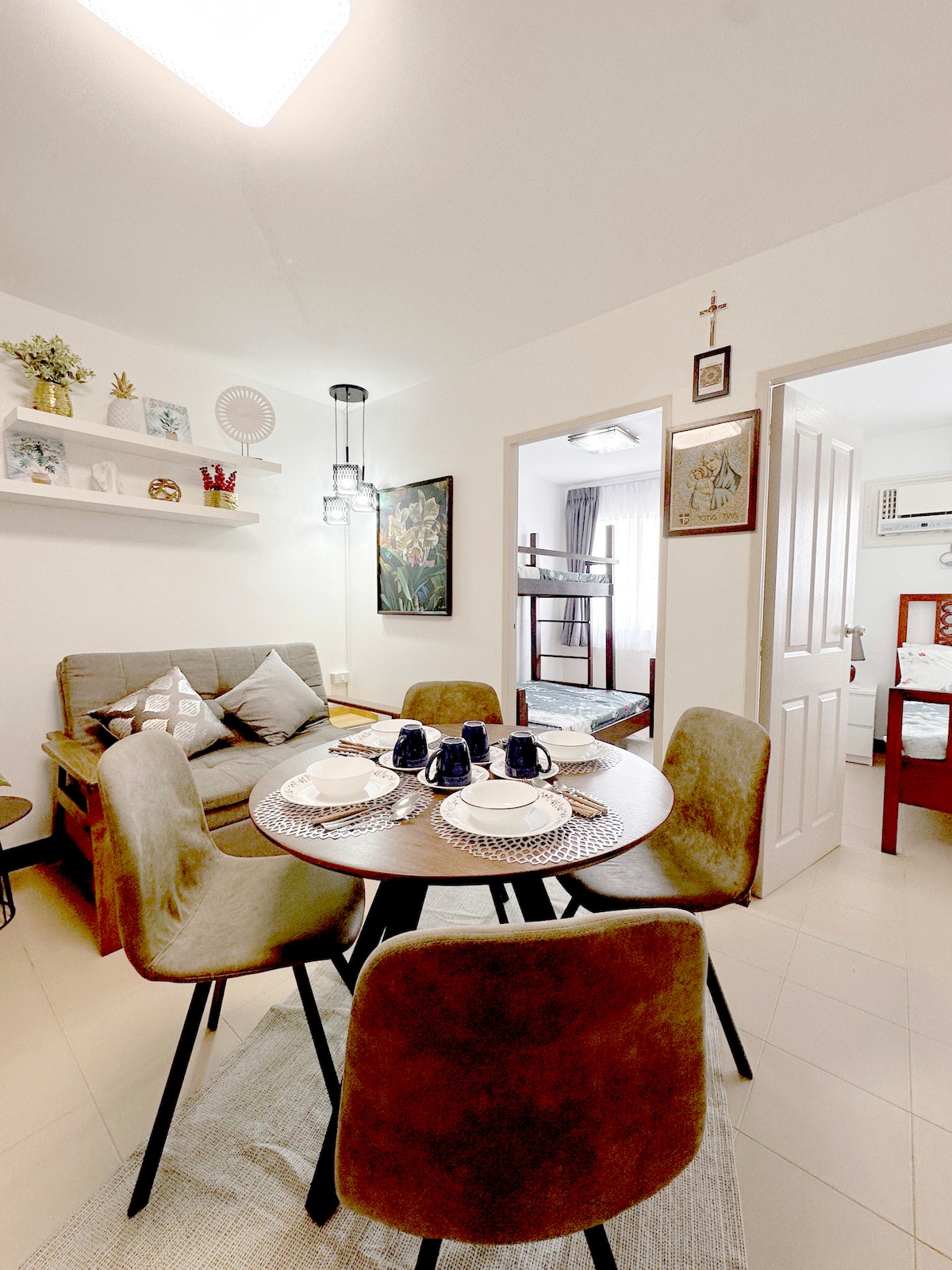 A dining area features a round table set for four, complemented by modern upholstered chairs. A cozy seating arrangement is visible nearby, with decorative items displayed on a shelf above. Doorways lead to the bedrooms and a window allows natural light to enter.