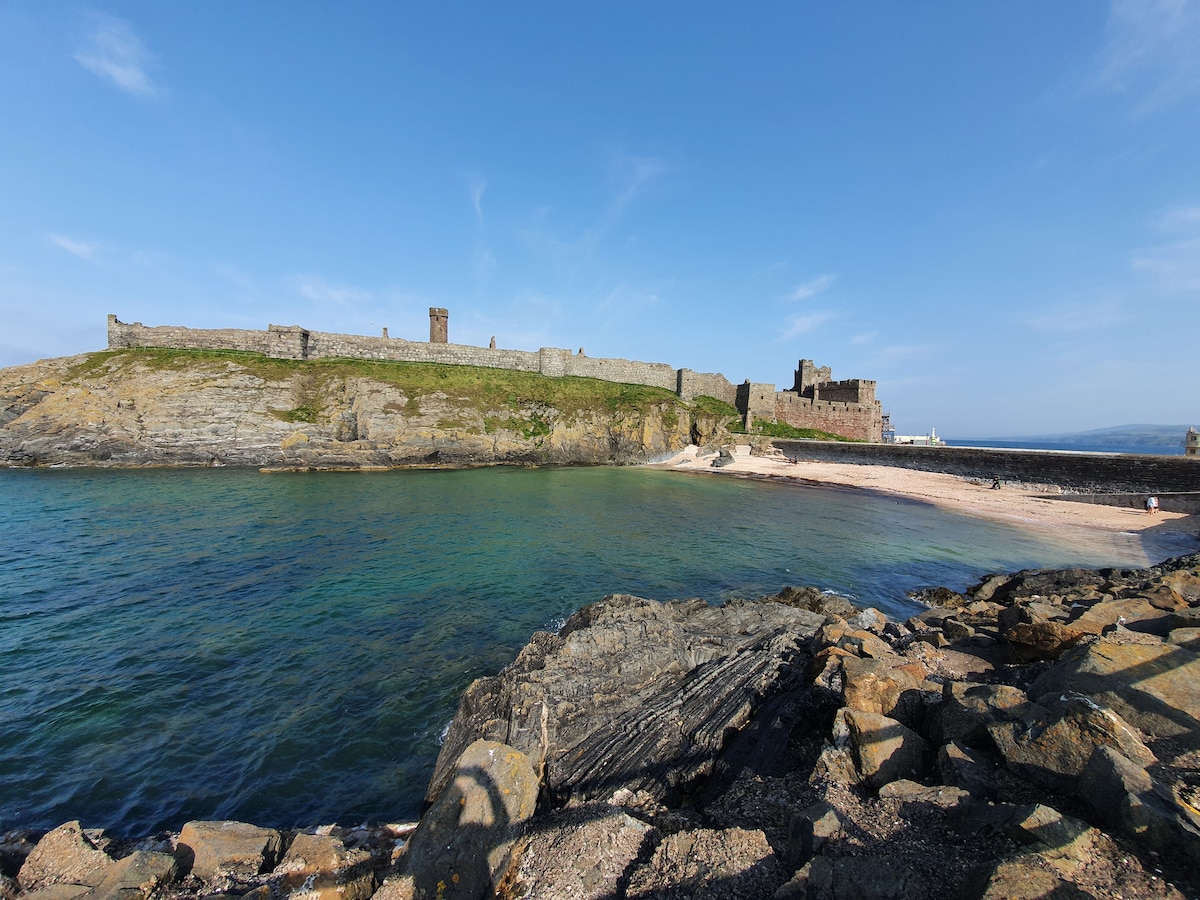 A coastal landscape features an ancient fortress perched on a rocky elevation, overlooking clear blue waters. The shoreline curves gently, highlighting sandy areas, while the distant hills provide a lush backdrop under a bright sky.