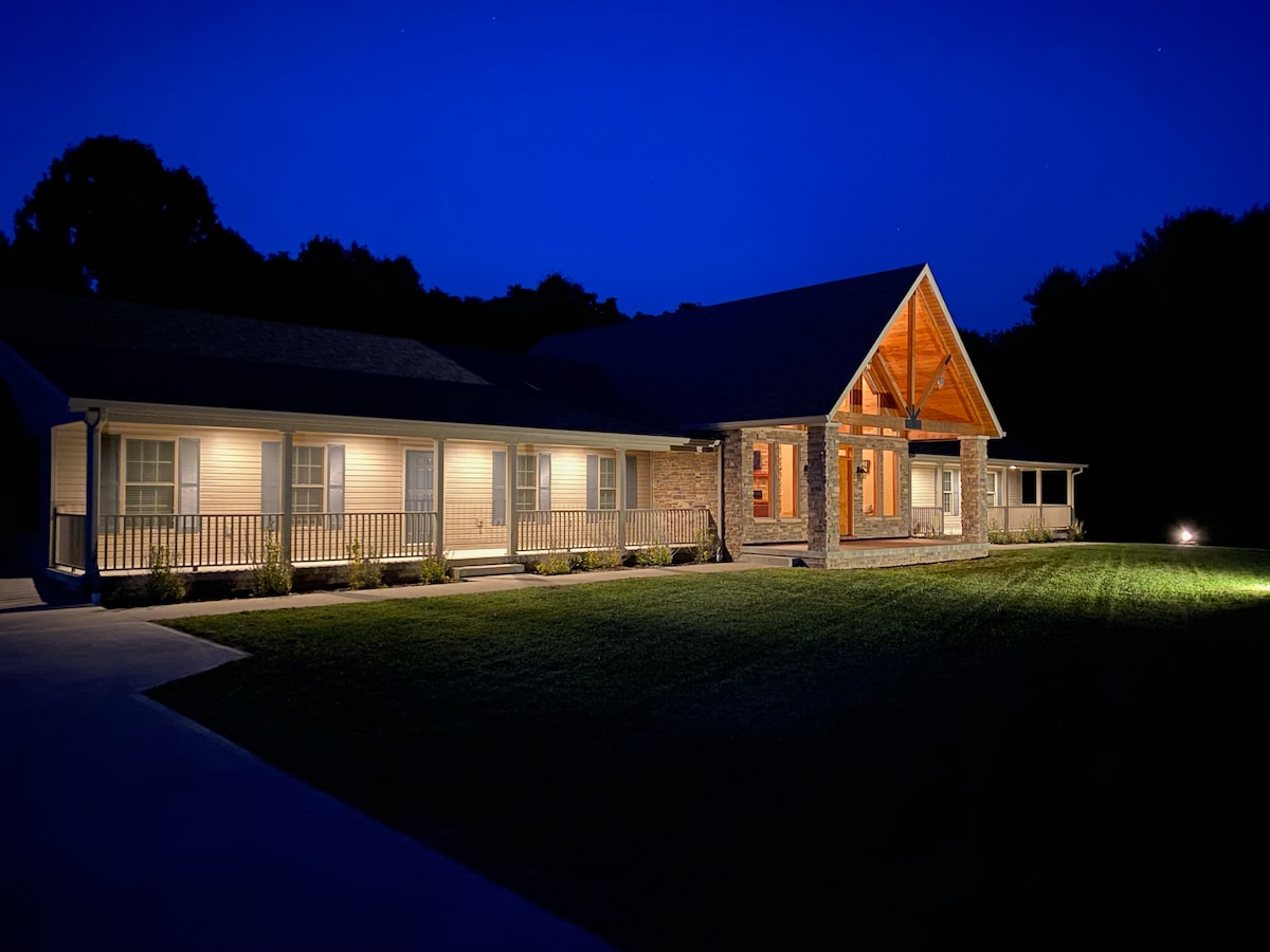 The exterior of the property is illuminated softly at night, showcasing its spacious design and multiple entries. A prominent wooden feature highlights the entryway, while landscaped grounds with carefully positioned lighting enhance visibility.