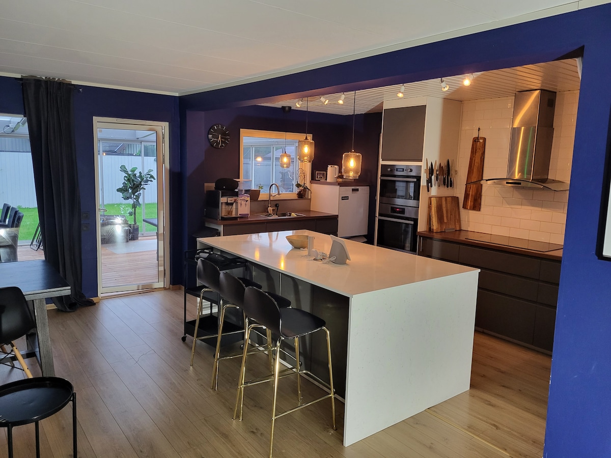 A modern kitchen features an island with a white countertop and seating for four. Warm lighting from pendant fixtures enhances the space. Dark cabinetry and stainless steel appliances are present, creating a functional area. A doorway leads to an outdoor space visible in the background.