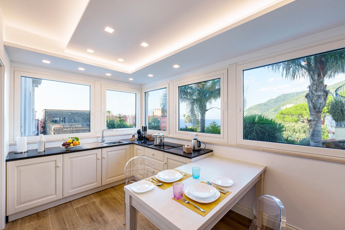 A light-filled kitchen is presented, featuring modern cabinetry and a countertop overlooking scenic views through large windows. A dining table is set with plates and colorful glasses, ready for a meal. Tropical plants and hills are visible outside, enhancing the harmonious environment.