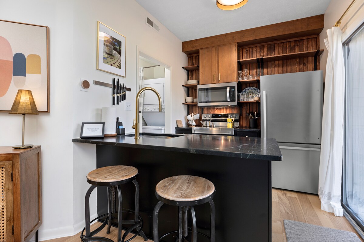 The kitchen area features a dark countertop paired with light wooden cabinetry. Stools are positioned at a compact island. Open shelves display glassware and decor. A modern refrigerator and microwave are included, enhancing functionality. A warm glow is provided by a hanging light fixture.