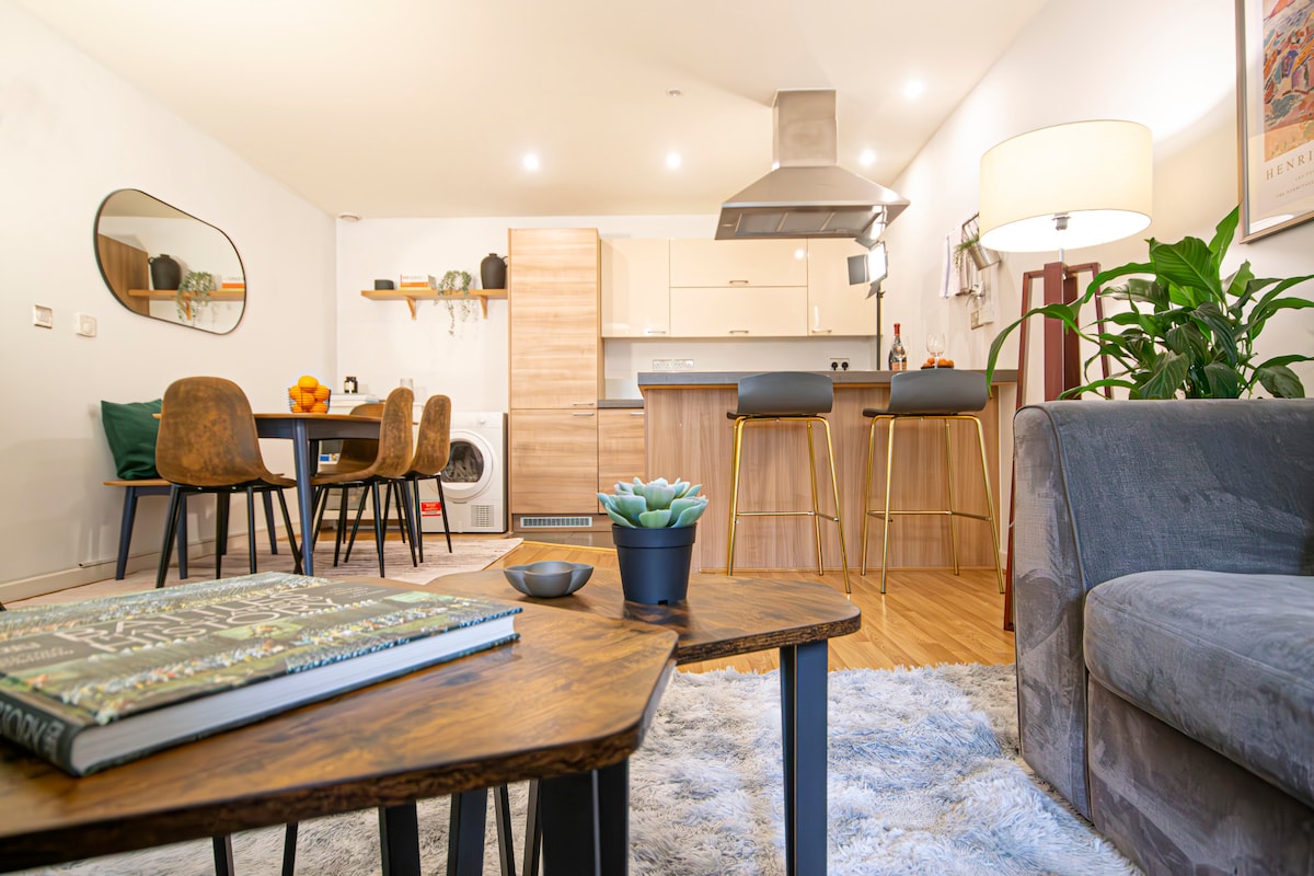 A thoughtfully arranged living area features a plush sofa paired with a stylish coffee table on a soft rug. A modern kitchen is visible in the background, complete with wooden cabinetry and contemporary bar stools around a sleek counter. Decorative plants and items add a welcoming touch.