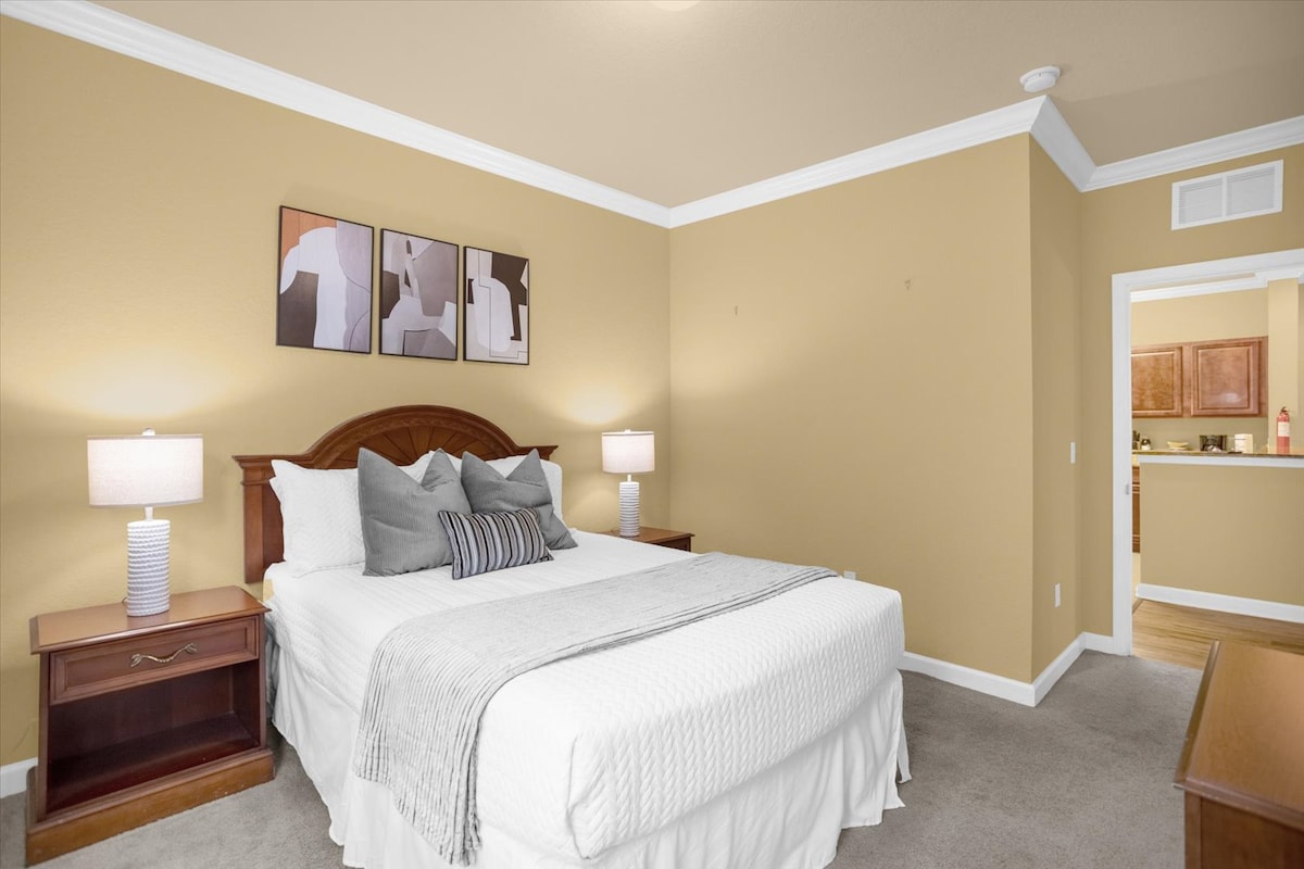 A spacious bedroom features a large bed with decorative pillows, flanked by nightstands with lamps. Artwork in neutral tones adorns the wall above the headboard. The room is painted in a warm golden hue, and a doorway leads towards a kitchen area.