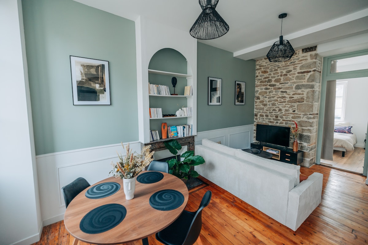 A bright living area features a soft, light-colored sofa and a round dining table set for four. Decorative shelves hold books, and greenery enhances the space. Exposed stone walls add character, while modern light fixtures complete the inviting atmosphere.