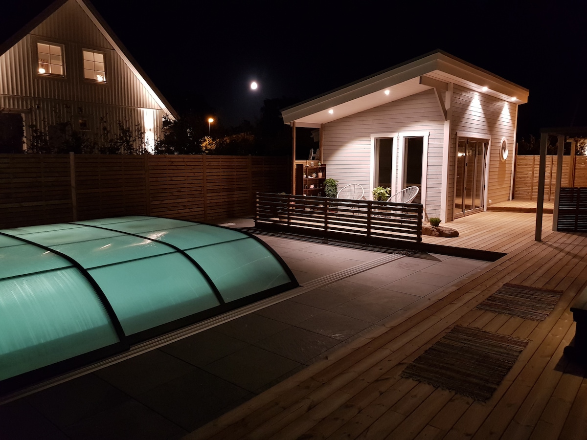 The illuminated exterior of the house showcases a modern design, featuring large windows and a patio area. A covered pool glows under the night sky, with moonlight reflecting off the water. Outdoor seating is available on the wooden deck, enhancing the tranquil ambiance.