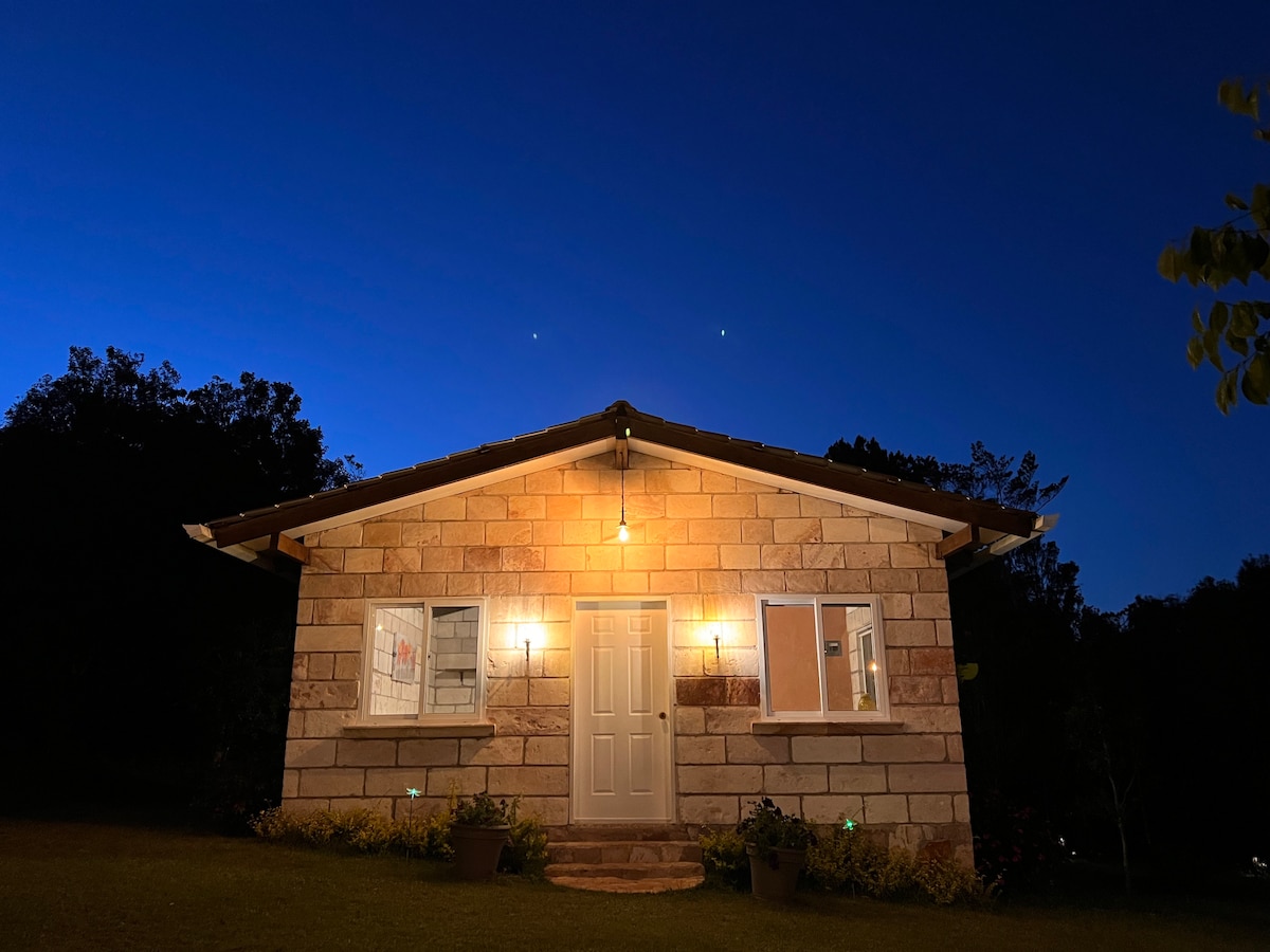 A cozy cabin stands under a twilight sky, featuring a warm glow from exterior lights. The stone façade is complemented by framed windows, while a welcoming door is centrally positioned. Surrounding greenery enhances the setting’s natural charm.