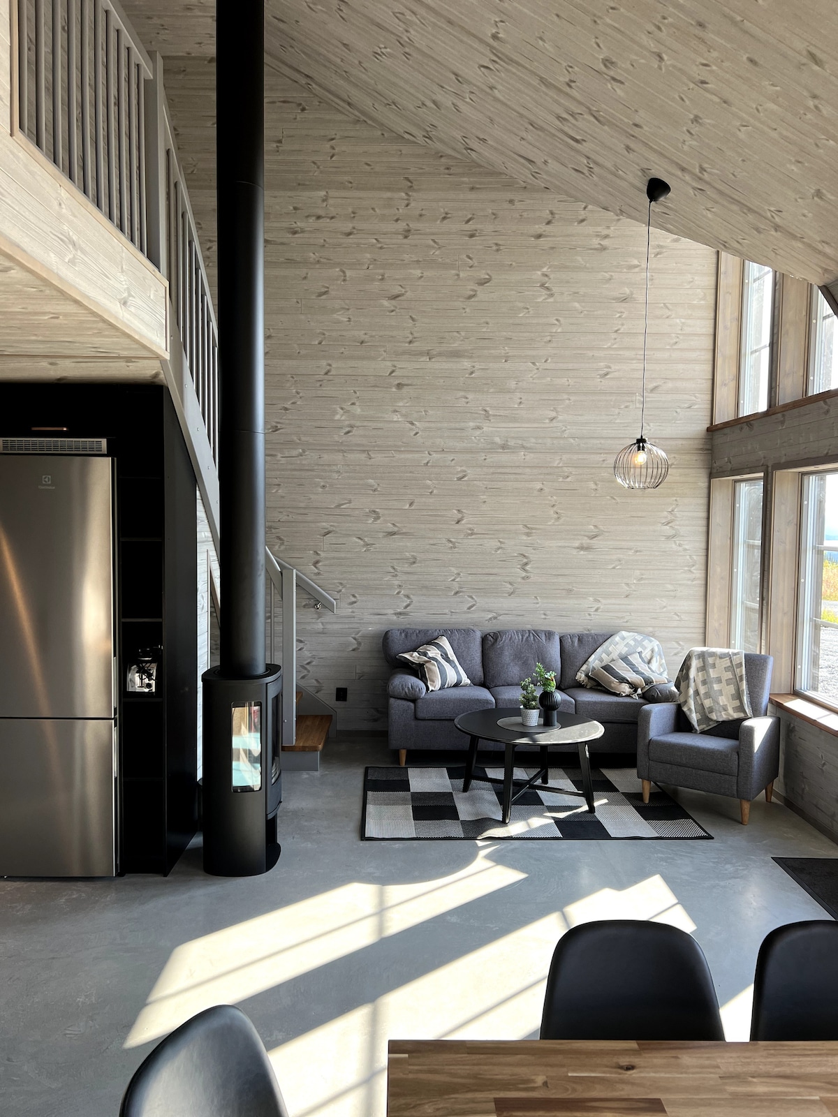 A bright living area features large windows allowing natural light to fill the space. A cozy seating arrangement of a sofa and chairs is complemented by a coffee table. A black wood-burning stove stands prominently beside the sleek kitchen area with modern appliances.