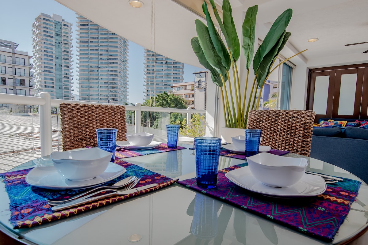 A dining area is presented with a glass table set for four, featuring white dishware and blue glasses. A vibrant, patterned table runner adds a touch of color. Lush green plants are positioned nearby, with large windows showcasing city views in the background.