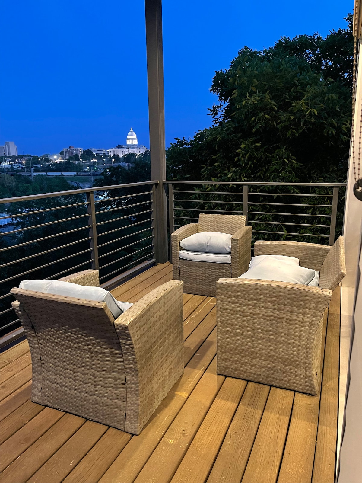 A cozy outdoor seating area is showcased on the deck, featuring two woven armchairs with soft cushions. The space overlooks the city, offering views of the Little Rock Capitol, with a twilight sky adding a serene backdrop.