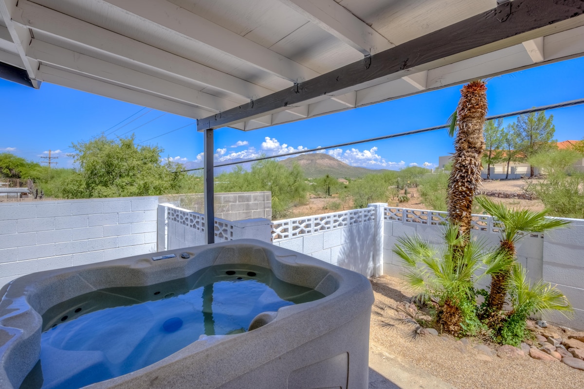 A hot tub is positioned under a covered area, surrounded by a patio. Lush palm trees and desert vegetation are visible beyond a low wall, offering views of the distant mountain range and bright blue sky.