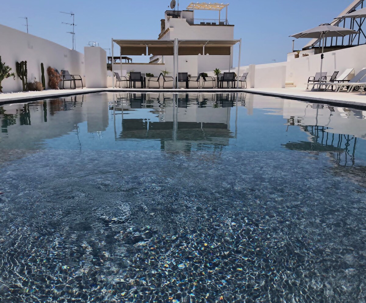 The image showcases a sleek outdoor infinity pool reflecting the clear blue sky. Comfortable sun loungers are arranged around the pool area, complemented by shaded seating under umbrellas. The surrounding white walls enhance the bright and airy atmosphere.