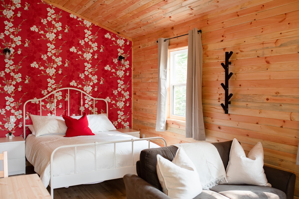 A cozy bedroom features a white metal bed with a red accent pillow and is framed by floral red wallpaper. Natural wood paneling complements the space, while a grey sofa with white cushions is positioned nearby. Soft light enters through a window dressed with light curtains.