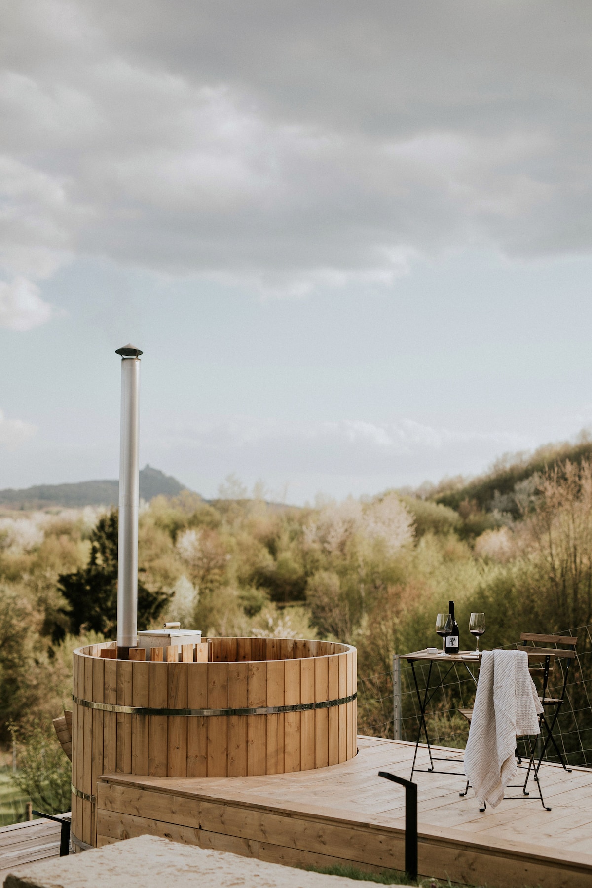 A wooden hot tub is situated on a spacious deck, accompanied by a small table and two chairs. A bottle and glasses are placed on the table. Lush greenery surrounds the area, providing a serene backdrop, with hills visible in the distance under a cloudy sky.