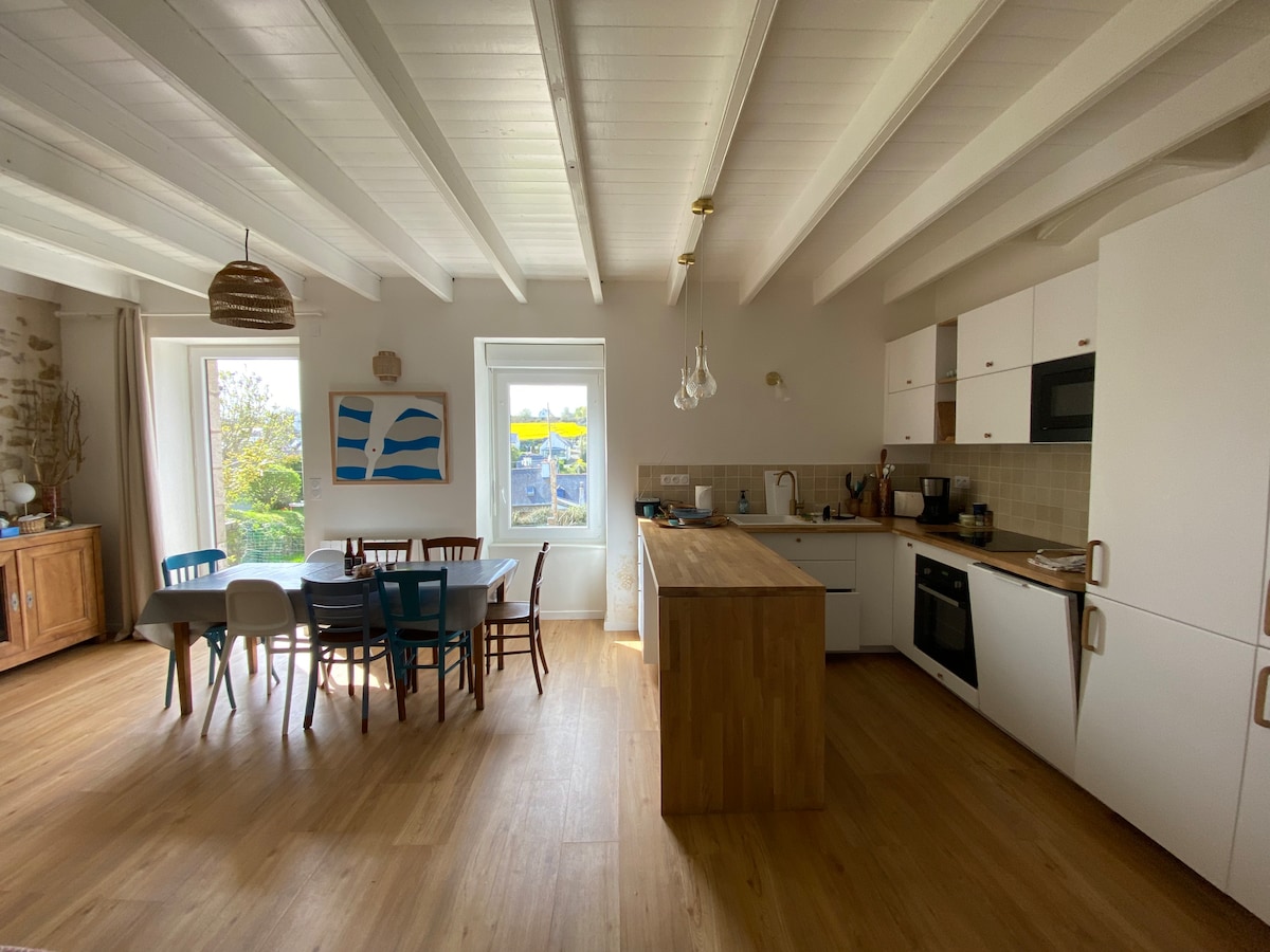 A functional kitchen and dining area is displayed, featuring wooden flooring and white-beamed ceilings. A large wooden island is centered, surrounded by dining chairs in various colors. A window provides natural light, revealing a glimpse of greenery outside.