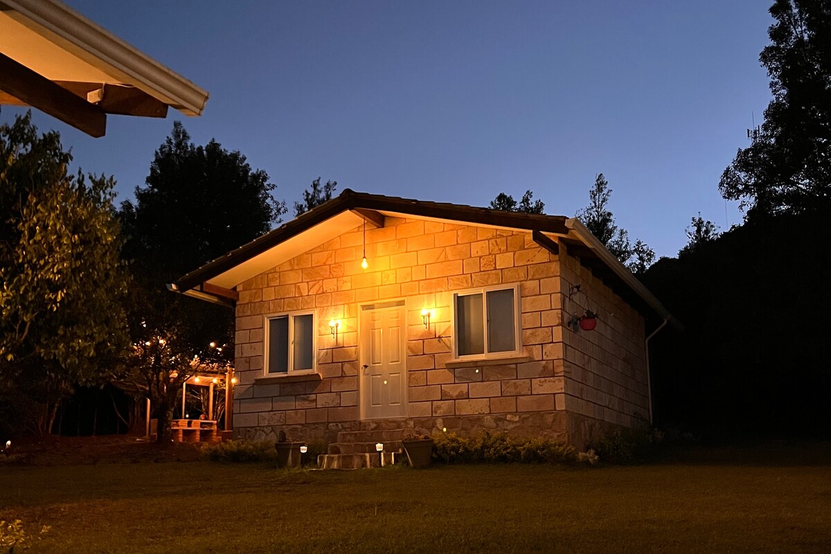 A charming cabin is illuminated against a twilight sky. Light emanates from two wall-mounted sconces flanking the front door, highlighting the stone facade. Surrounding greenery adds a natural touch, while the silhouette of trees stands in the background, creating a serene atmosphere.