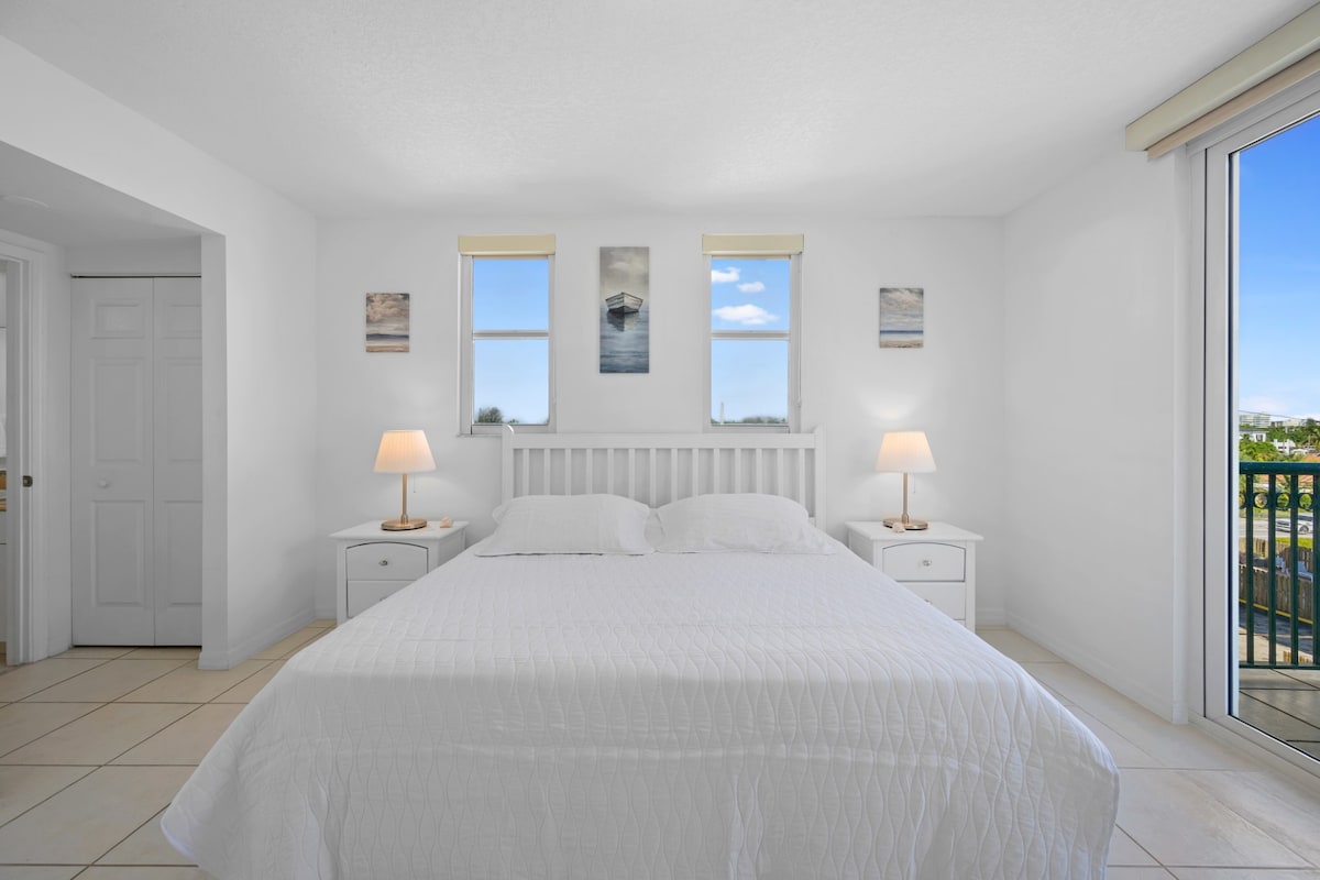A bright and airy bedroom features a spacious bed with a white quilted coverlet, flanked by two bedside tables with lamps. Natural light pours in from two windows, and a sliding glass door leads to a private balcony with a view.