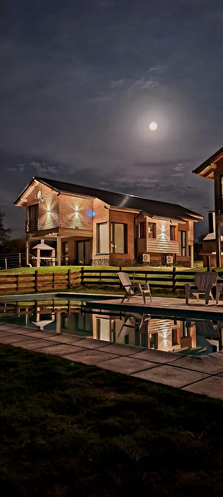 A modern country house is seen at night, illuminated by soft exterior lighting and the moon overhead. Reflection in the nearby swimming pool highlights the house's architectural features. An expansive lawn and fence frame the property, adding to the serene environment.