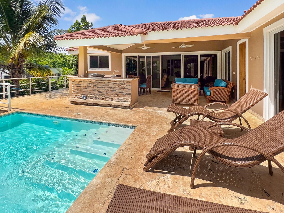 A sun-drenched patio area features a sparkling pool surrounded by comfortable lounge chairs. A barbecue station is visible, alongside large glass doors leading to the interior. Lush greenery provides a serene backdrop against a clear blue sky.