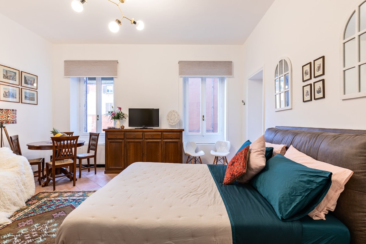 A spacious room is featured, equipped with a comfortable bed adorned with decorative pillows. A small dining table with chairs is positioned beside a wooden cabinet and a television. Natural light enters through windows, creating a bright atmosphere complemented by neutral-toned decor.