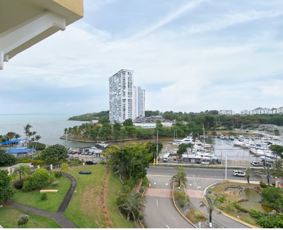 A panoramic view of the coastline is captured, featuring a marina with several boats docked. The surrounding greenery enhances the natural setting, while modern buildings rise in the background. The image highlights a blend of water, greenery, and urban elements in the landscape.