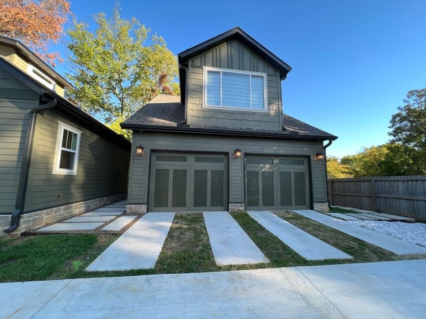 The exterior of the studio apartment is shown, featuring a two-story structure with dark siding and a pitched roof. Large, double garage doors are visible, framed by a concrete driveway and bordered by neatly maintained grass. A gravel area complements the landscaping on the side.