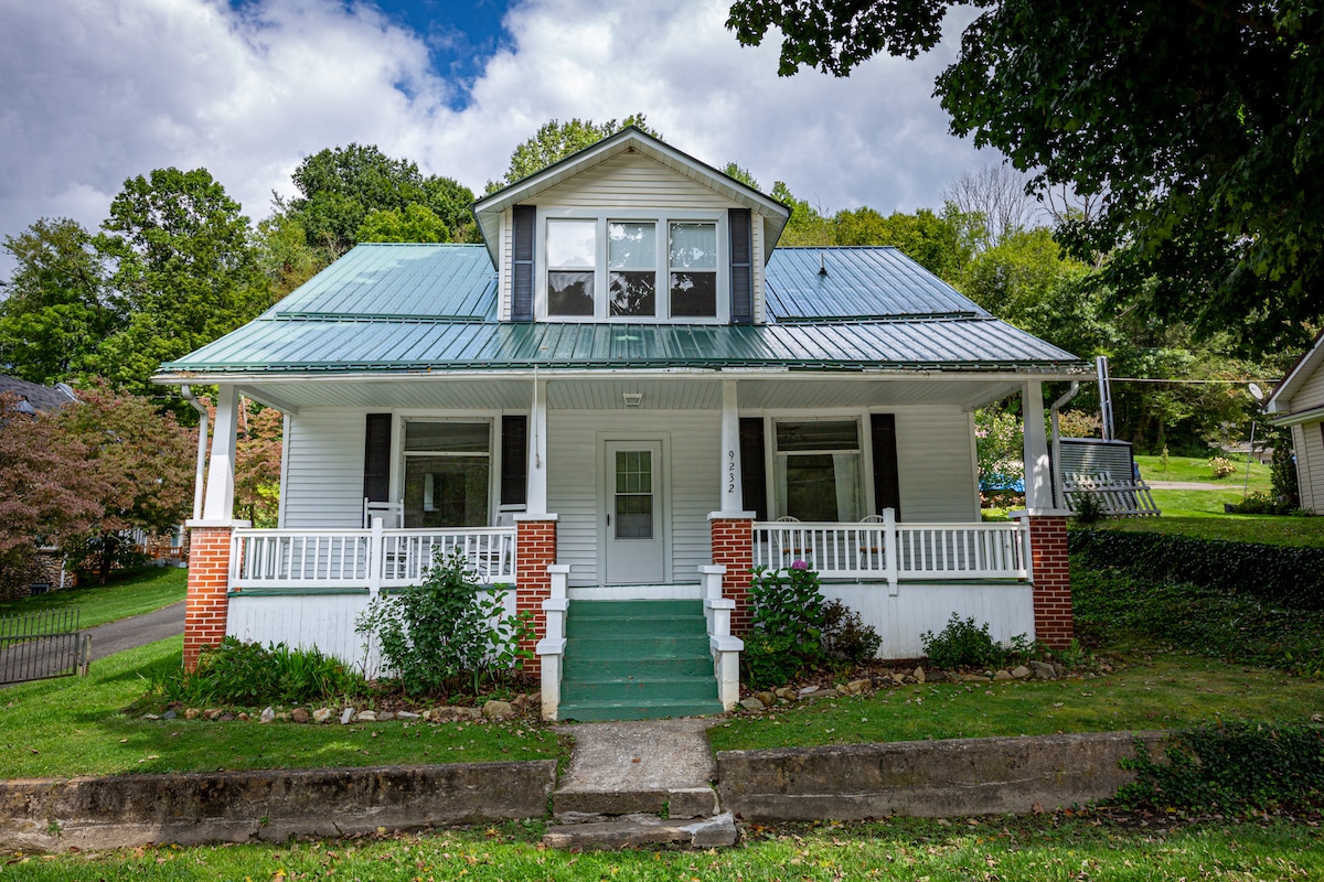 The charming house features a covered front porch with white railings and green steps. A metal roof reflects natural light, while large windows invite ample daylight. Surrounding greenery complements the inviting exterior, creating a welcoming entrance to the home.