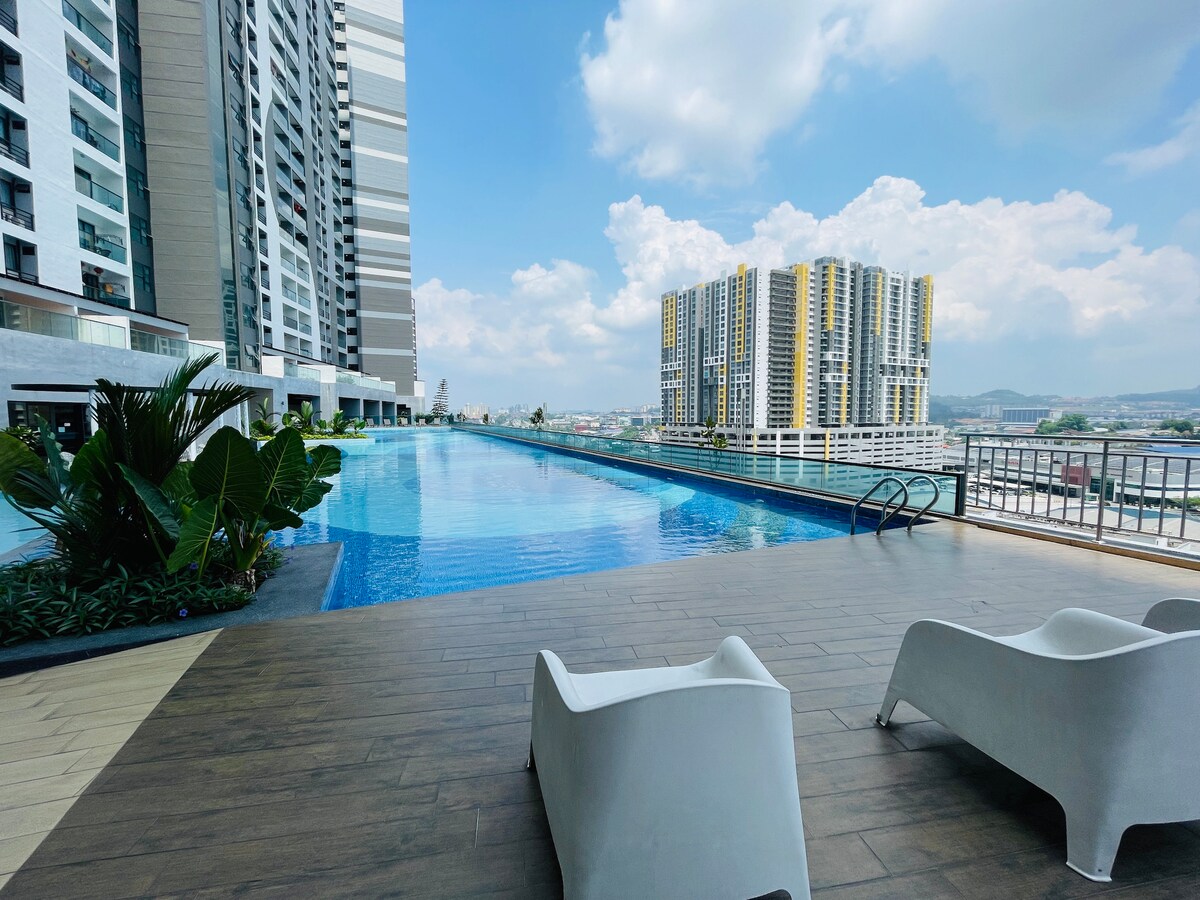 The rooftop swimming pool area is featured, showcasing a long infinity pool that reflects the sky. Comfortable white loungers are placed on the wooden deck beside the pool, with greenery surrounding the area. The expansive view of nearby buildings and the horizon is visible.