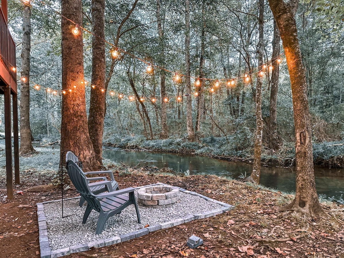 A serene outdoor area features two comfortable chairs positioned near a small fire pit, bordered by circular stones. Warm string lights are draped between trees, casting a soft glow over the nearby flowing creek, surrounded by lush greenery.