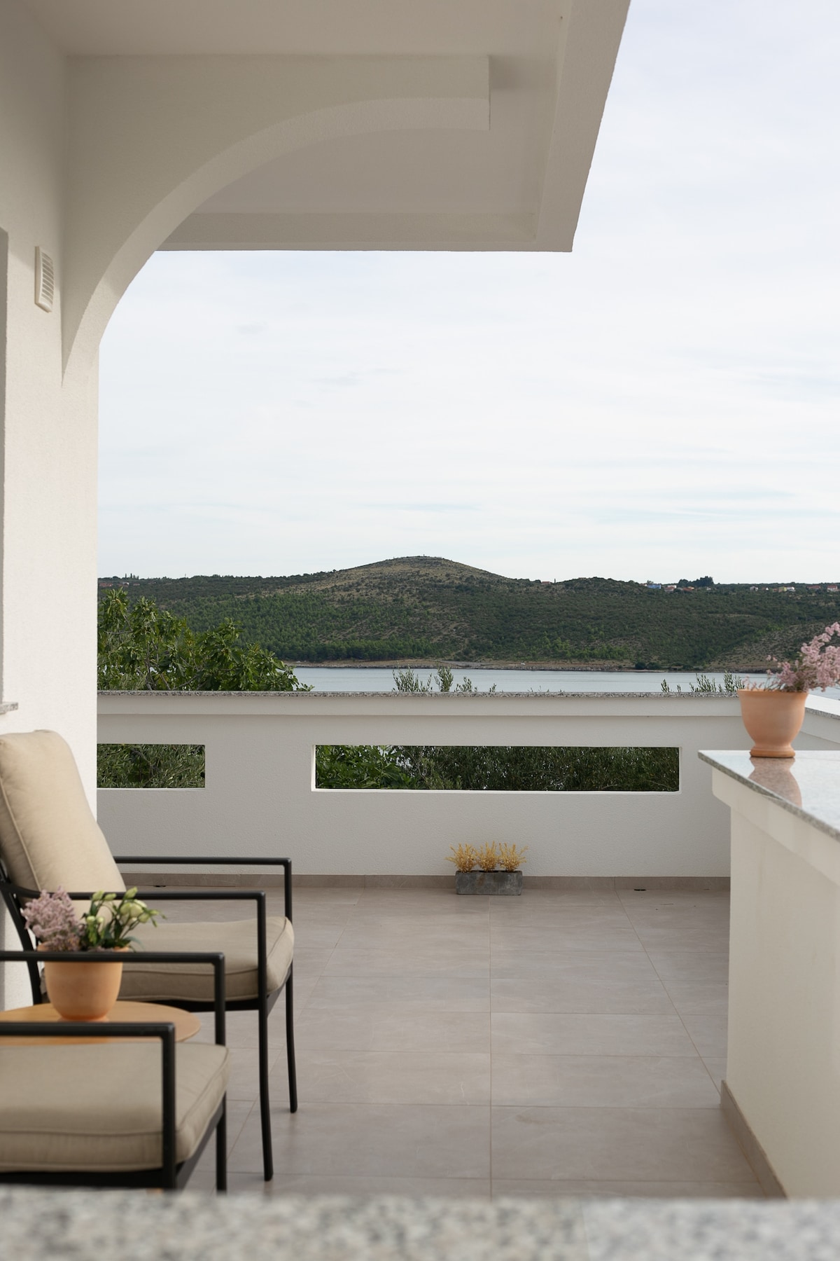 A spacious balcony is shown, featuring two comfortable chairs positioned beside a small table. Potted flowers add a touch of greenery, while a serene view of the landscape and water can be seen in the background, enhancing the outdoor relaxation space.