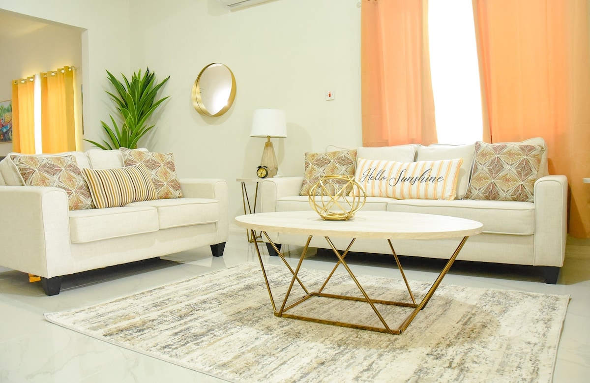 A spacious living area is presented with light-colored sofas adorned with decorative cushions. A circular coffee table is positioned at the center, complemented by a decorative bowl. Natural light filters through sheer orange curtains, enhancing the welcoming ambiance.