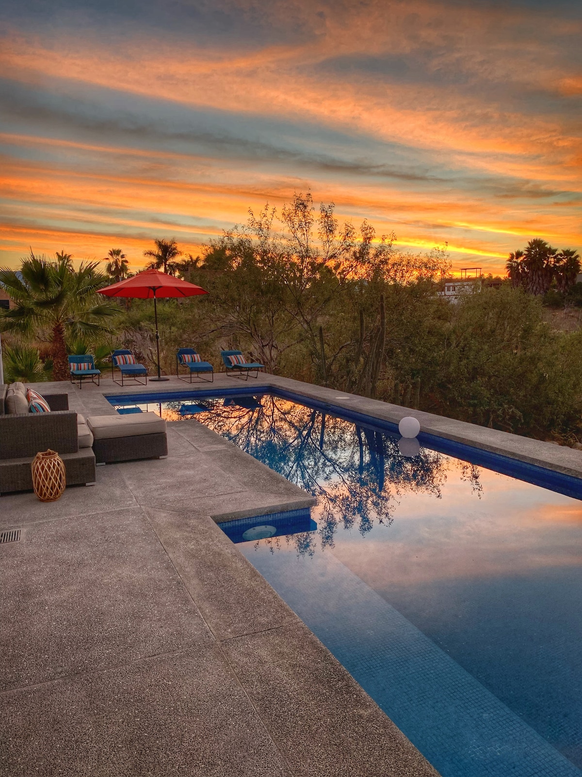 A serene outdoor area showcases a tranquil salt-water pool reflecting the colorful sunset. Comfortable seating arrangements surround the pool, with lounge chairs and a large umbrella providing shade. Lush greenery frames the space, enhancing the peaceful ambiance.