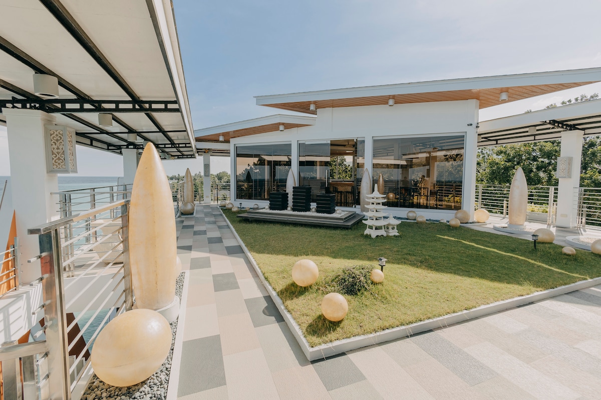 An outdoor space features a well-manicured lawn, surrounded by modern architecture. Lounge areas with dark furniture and decorative elements are visible. Sculptural features and smooth stones add visual interest, while expansive glass walls invite natural light into the building.