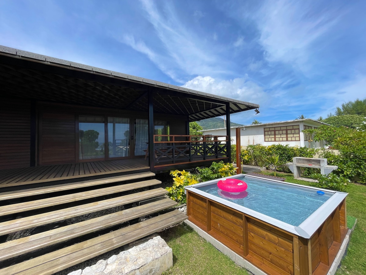 A bungalow is shown with a spacious wooden deck leading to the entrance. A small pool is visible in the foreground, featuring a bright pink float. Lush greenery surrounds the area, with a clear sky overhead.