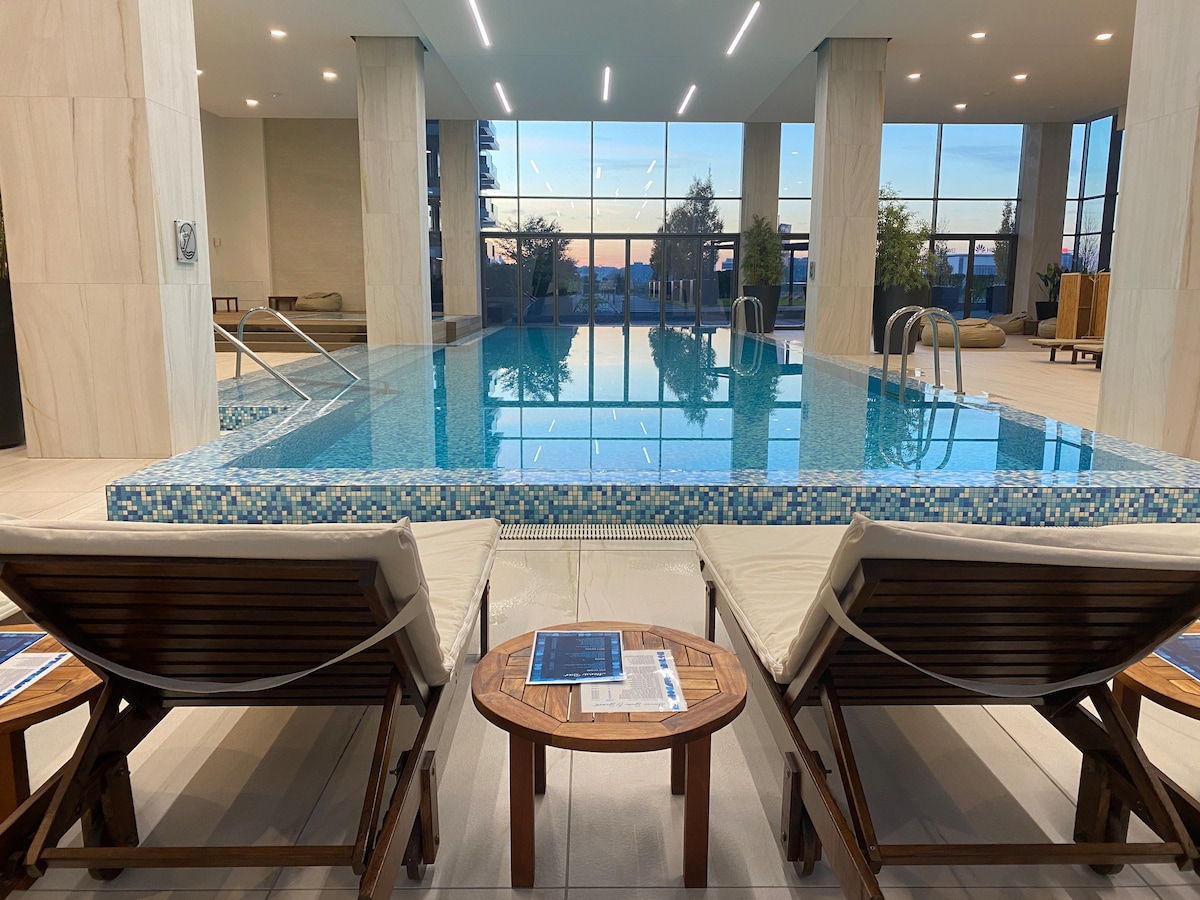 An indoor pool area is highlighted by a tranquil water feature, surrounded by modern seating arrangements. Two sun loungers and a small wooden table are positioned by the water's edge, with large windows offering views of the surrounding area and allowing natural light to fill the space.