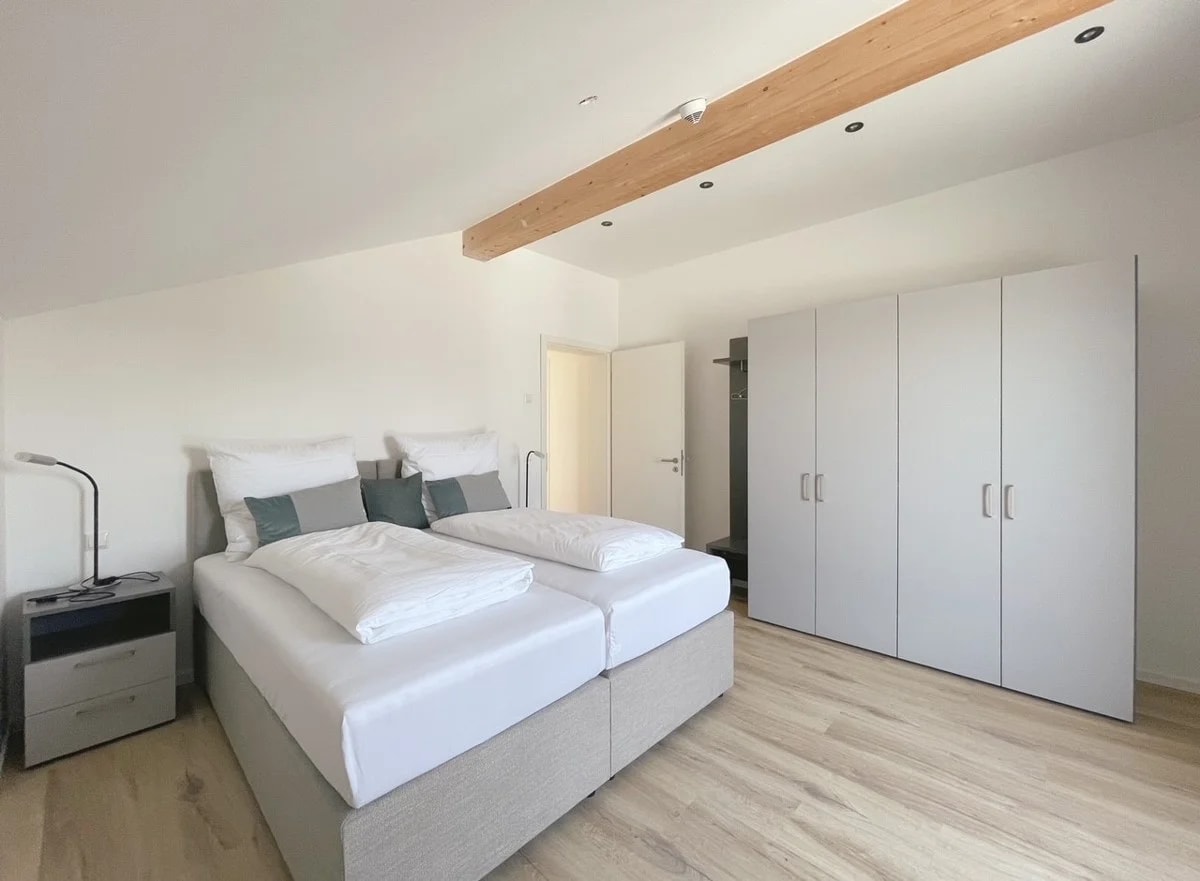 A bright bedroom features a spacious boxspring bed, which can be separated into two single beds. A modern wardrobe stands against the wall, complemented by a sleek nightstand with a reading lamp. Natural light filters softly through the angled ceiling.