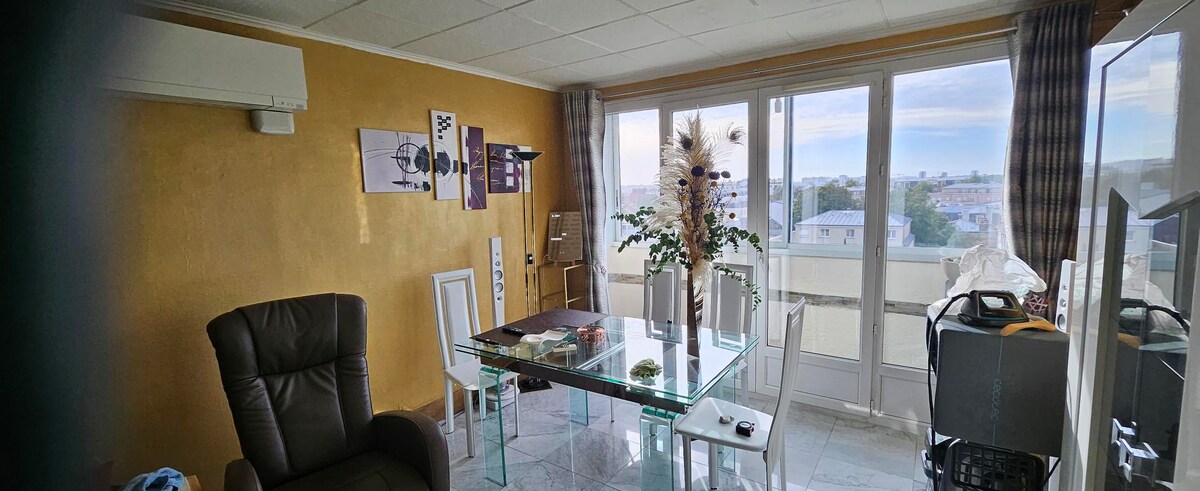 A bright living area features a glass dining table surrounded by chairs. Large windows offer views of the outside, while soft natural light illuminates the space. Decorative elements, including a vase with plants, add character to the room, complemented by stylish artwork on the walls.