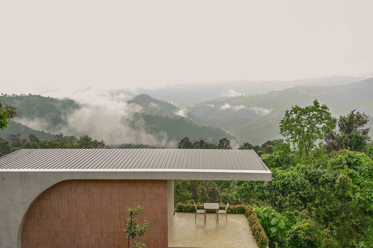 The image showcases a modern villa set against a backdrop of misty mountains. A flat-roofed structure features a large terrace with a simple seating arrangement, surrounded by lush green foliage, creating a seamless connection to the serene natural landscape.