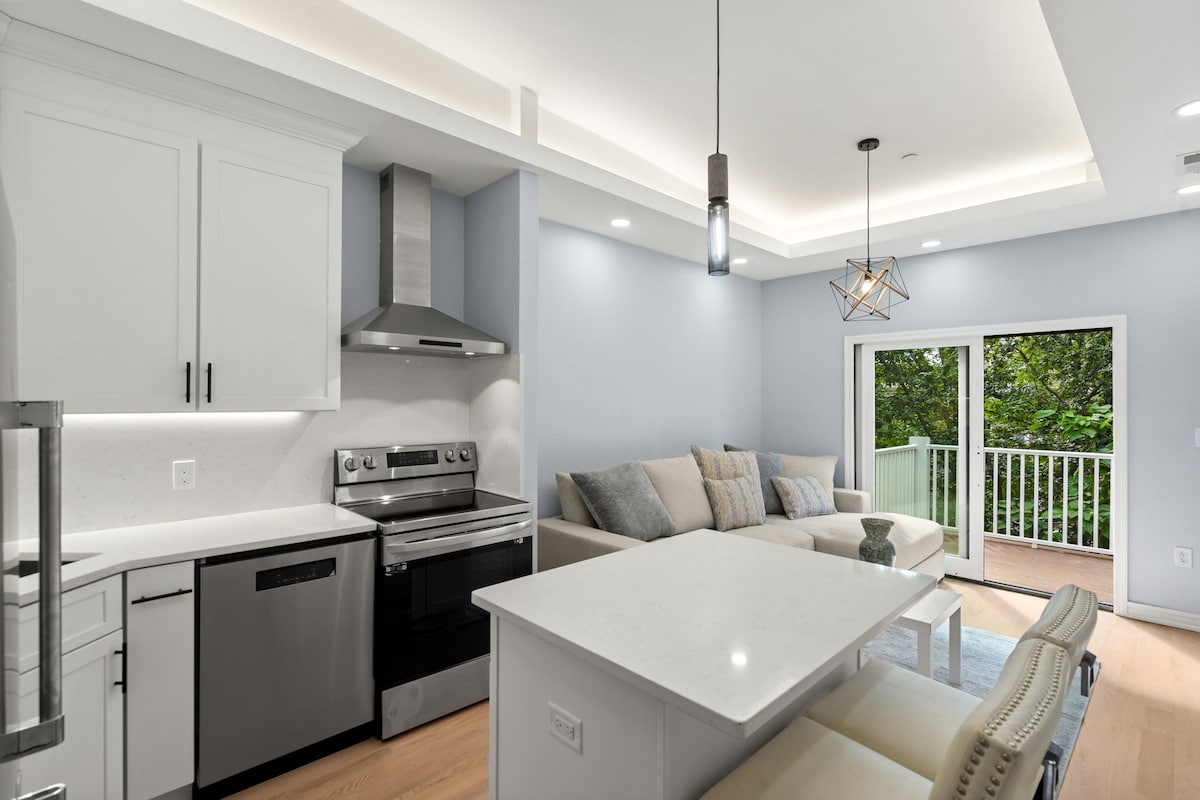 A modern kitchen and living area feature sleek appliances, light cabinetry, and a cozy sectional sofa. A dining table with chairs offers ample seating. Large sliding doors open to the private balcony, allowing natural light and views of greenery to fill the space.