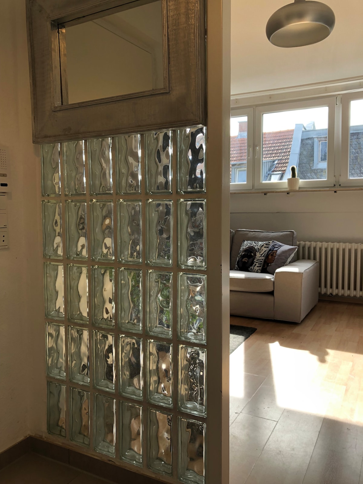 A glass block wall separates two spaces, allowing natural light to filter through. In the background, a cozy seating area is visible, featuring a light-colored sofa with decorative cushions. Large windows provide views of the outside, and wooden flooring adds warmth to the area.